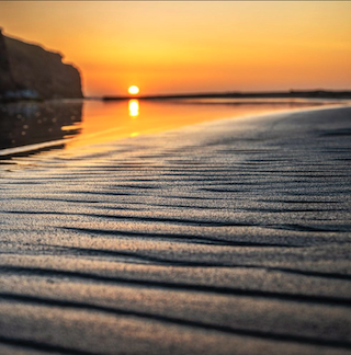 Close-up of sun-kissed beach sand with vibrant orange and yellow sunset reflections on the water, capturing the serene beauty of a coastal evening.